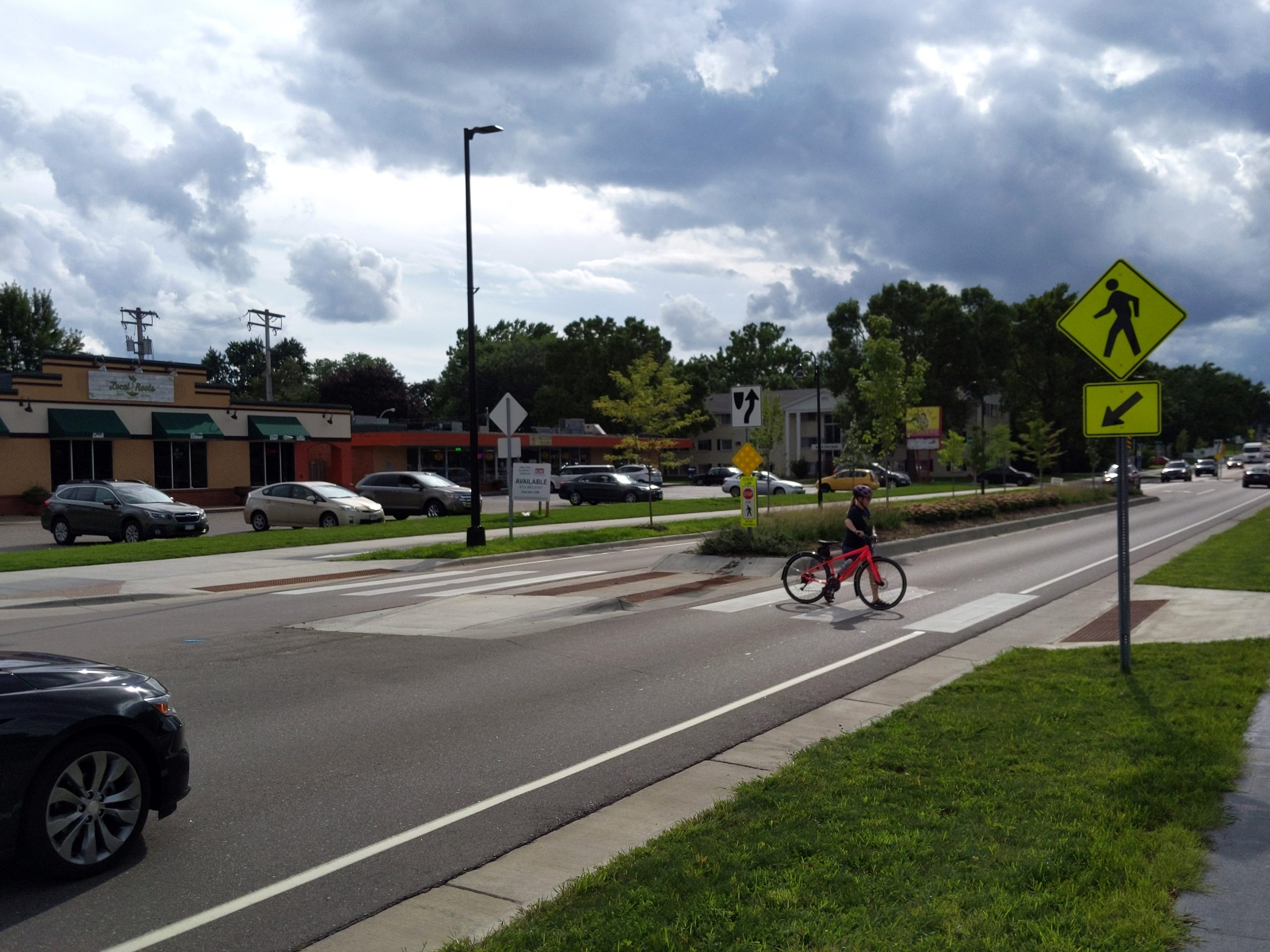 A pedestrian crosses at one of many pedestrian crossing islands on the new 66th Street. Adjacent to a major park, this section of road was previously 4 lanes with no medians, bike space, or greenery.